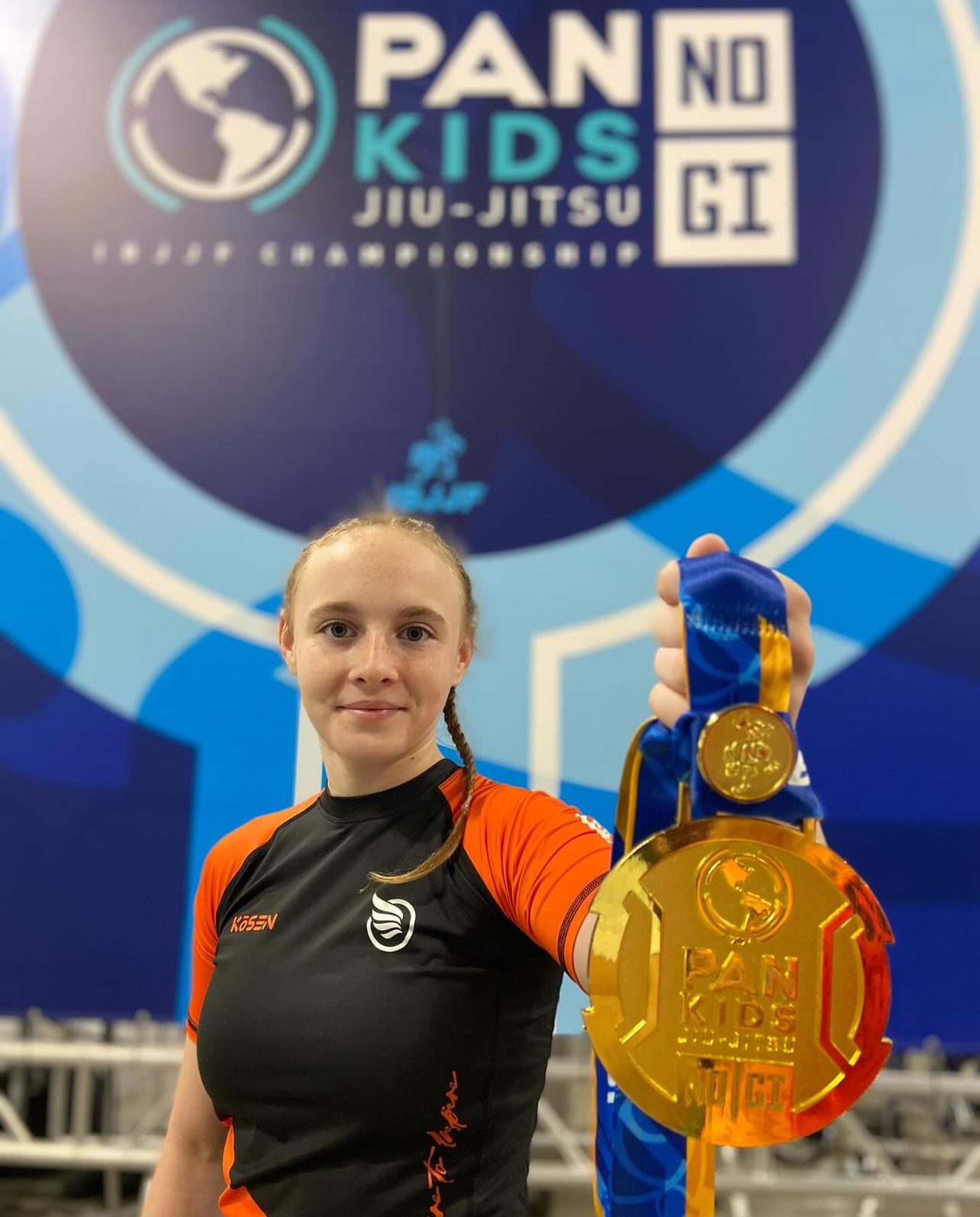 Fotografía de la medalla de oro tomada en el Pan Kids IBJJF No-Gi Championship 2024. La medalla lleva la identificación del Pan Kids IBJJF Championship. El fondo del evento PAN KIDS NO-GI IBJJF CHAMPIONSHIP es visible detrás.