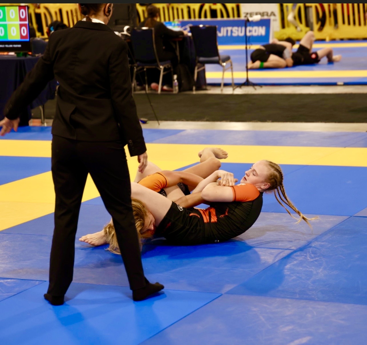 Imagen de competencia del cuadro del Pan Kids IBJJF No-Gi Championship 2024. Se muestra a la protagonista en posición de control superior. Entorno de competencia sancionado por la IBJJF, con la mesa de puntuación y los tapetes contiguos visibles al fondo.